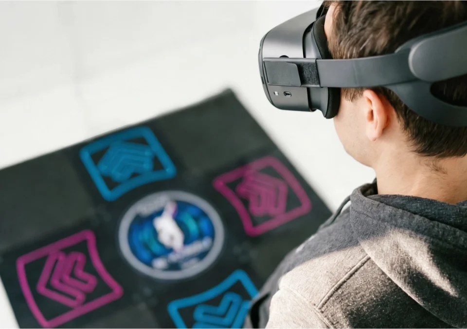 A person wearing a VR headset looking down at an L-TEK dance pad with neon blue and pink directional arrows — combining virtual reality and rhythm gaming