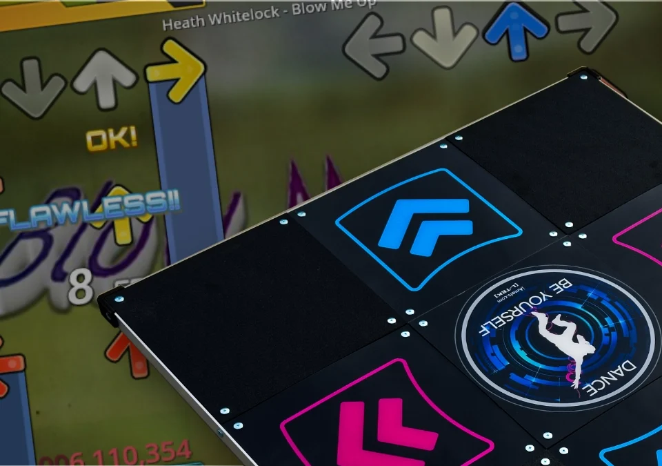 L-TEK Dance Pad with blue and pink directional arrows placed in front of a StepMania gameplay screen showing a "FLAWLESS!!" rating and score of 006,110,354 on the song "Blow Me Up" by Heath Whitelock