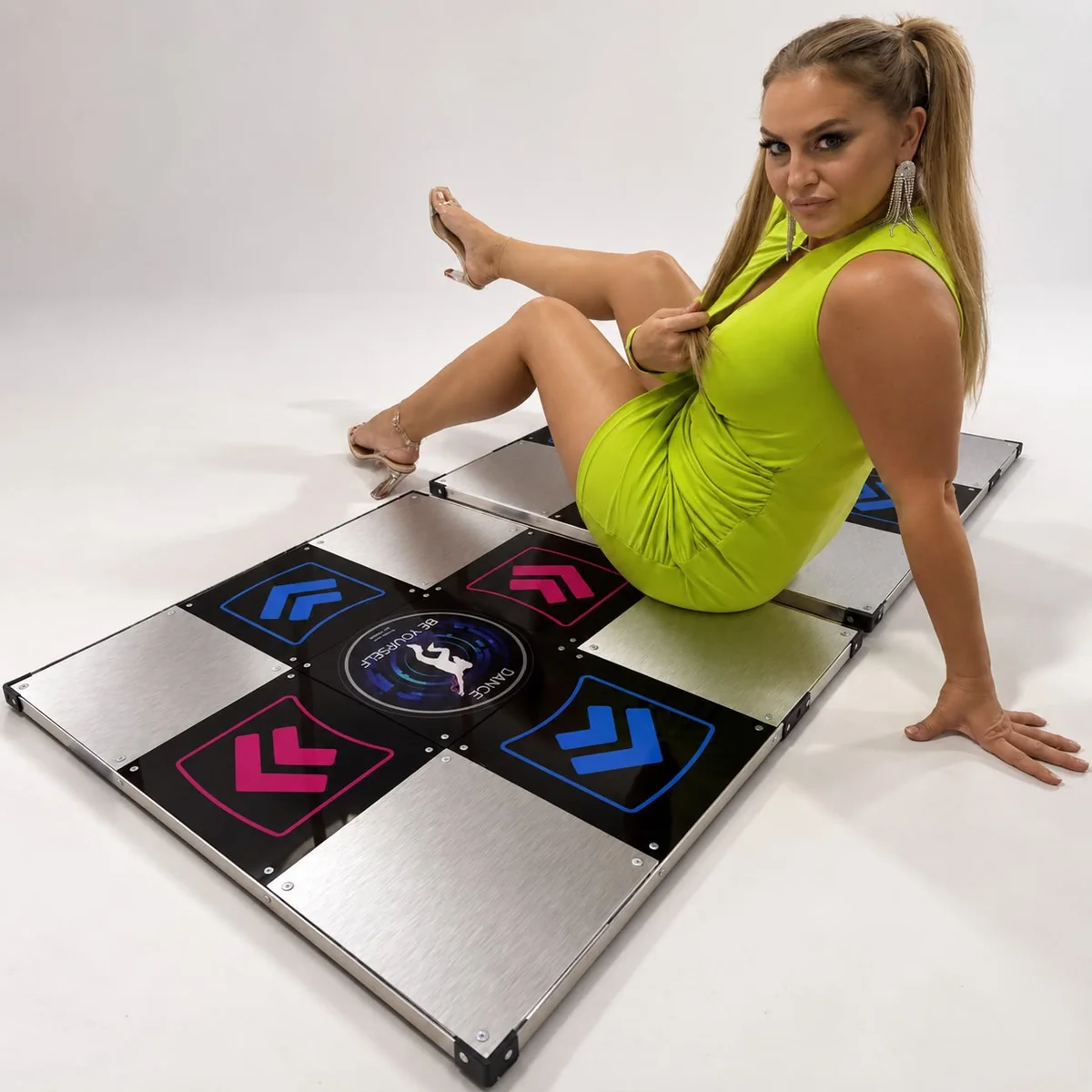 L-TEK メタルブラック ダンスパッド – ライフスタイル - Interactive Mats L-TEK ライム色のドレスを着た女性が、ブラッシュドメタル調のパネルとピンクとブルーの矢印パネルを備えた「L-TEK Metal Black」ハードダンスパッドの上に座っている